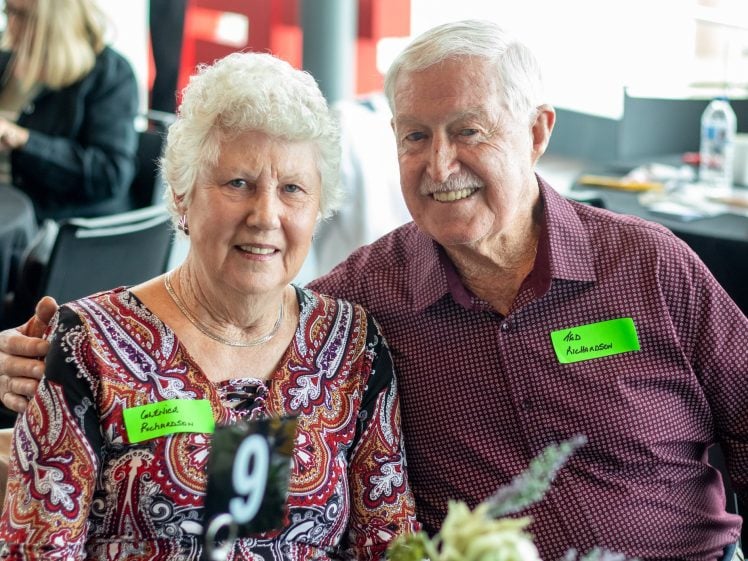 Image of An elderly woman and man, both smiling, sit closely together at a table with a black number 9 sign. They wear name badges and patterned shirts in a bright indoor setting with blurred people in the background.