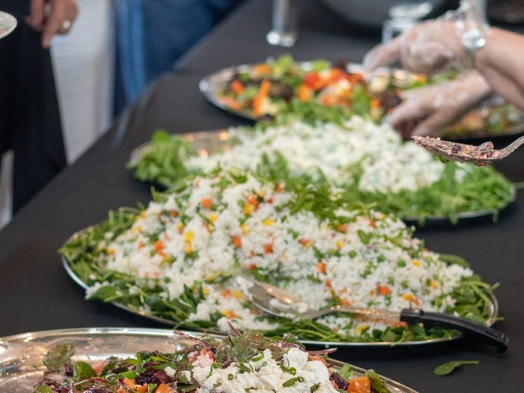 Image of A buffet table with large platters of colourful salads, including leafy greens and diced vegetables. People are helping themselves to food, and one person is using tongs while wearing gloves.