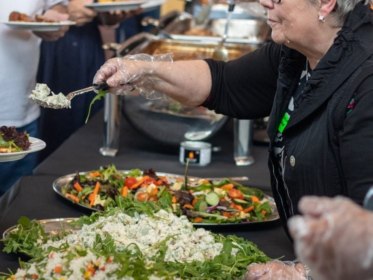 Image of A group of people serve themselves food from a buffet table filled with large trays of salad and rice dishes. One woman in the foreground, wearing gloves, is spooning salad onto a plate.