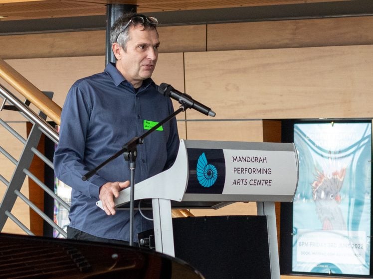 Image of A man in a blue shirt speaks into a microphone at a podium labelled Mandurah Performing Arts Centre. He is indoors, standing near a staircase, and posters are visible in the background.
