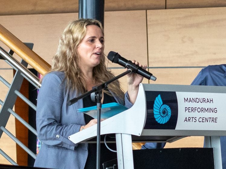 Image of A woman with long blonde hair speaks into a microphone at a lectern labelled Mandurah Performing Arts Centre. She holds papers and stands in front of a staircase indoors.