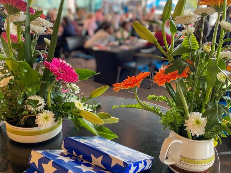 Image of Two vases with colourful flowers and three presents wrapped in blue paper with white stars sit on a round black table, with a blurred background of people gathered indoors.