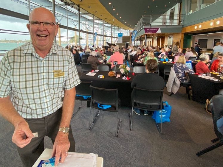 Image of A smiling man in glasses and a checked shirt stands at the front of a large, busy room filled with people seated at tables, suggesting a lively social or community event.