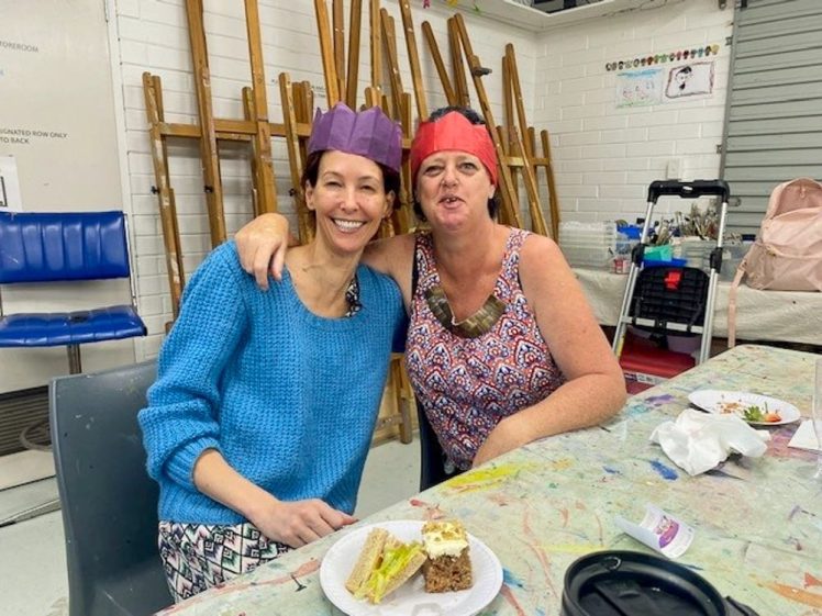 Image of Two women wearing colourful paper crowns sit together at a table, smiling and enjoying food. Art easels are in the background, and the table is scattered with plates, cake, and snacks in a brightly lit room.