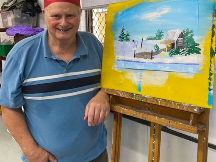 Image of A smiling man in a red paper crown stands next to an easel displaying his painting of a snowy cabin scene with trees, a jetty, and a blue sky, all framed by a yellow border.