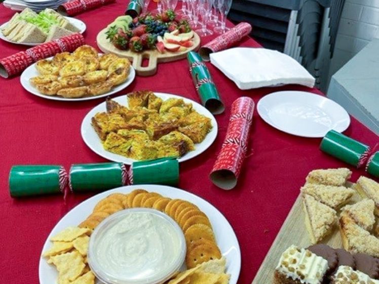 Image of A table with a red tablecloth is set with plates of crackers and dip, sliced cake, savoury pastries, biscuits, sandwiches, fruit, and bottles of orange squash and water. Stacked chairs and glasses are in the background.