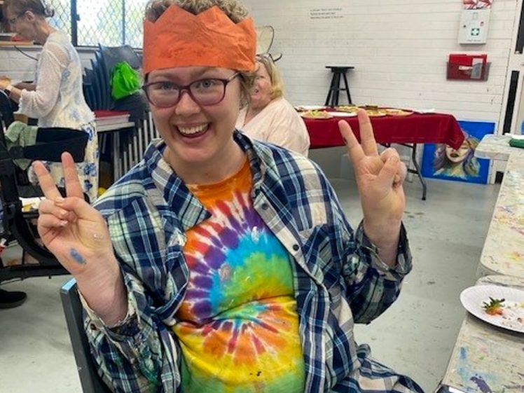 Image of A smiling person wearing glasses, a red paper crown, a tie-dye shirt, and a checked shirt gives a double peace sign. They are seated in an art studio with art supplies and other people in the background.