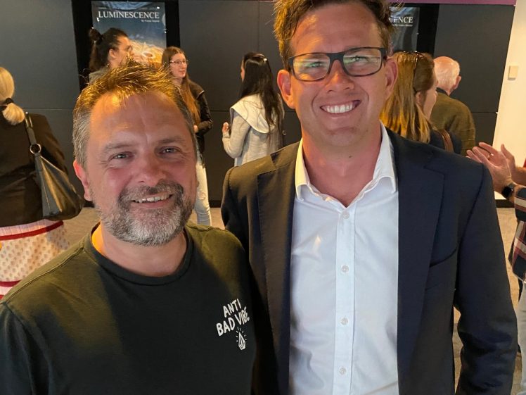 Image of Two men smiling and posing for a photo indoors. One is wearing a dark T-shirt, and the other is wearing glasses, a white shirt, and a dark suit. People and a sign reading MEZ Art Space are visible in the background.