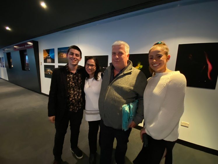 Image of Four people stand together indoors, smiling at the camera. Framed photographs are displayed on the wall behind them. The group appears to be at an art exhibition or gallery.