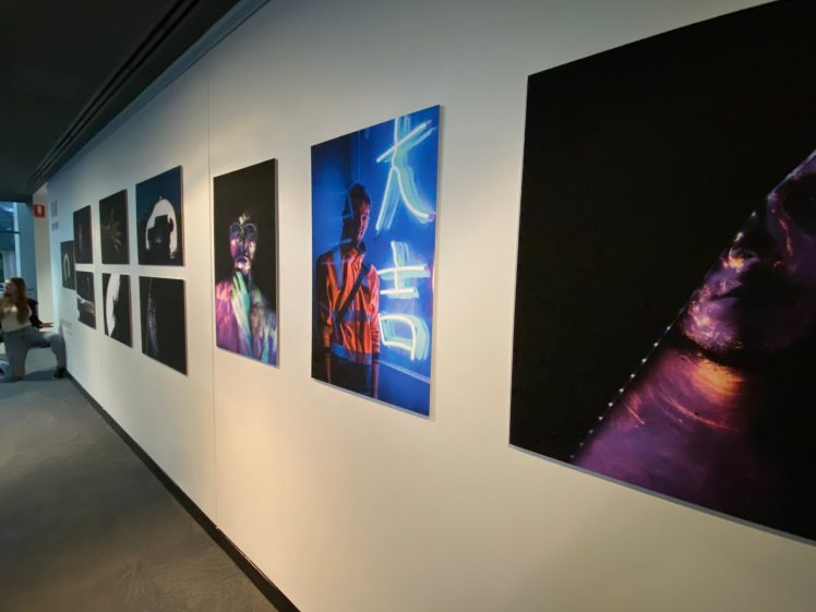 Image of A corridor art exhibition displays large, colourful, and moody photographs mounted on a white wall, featuring dramatic lighting and neon colours. A person is seen in the far left corner, mostly out of frame.