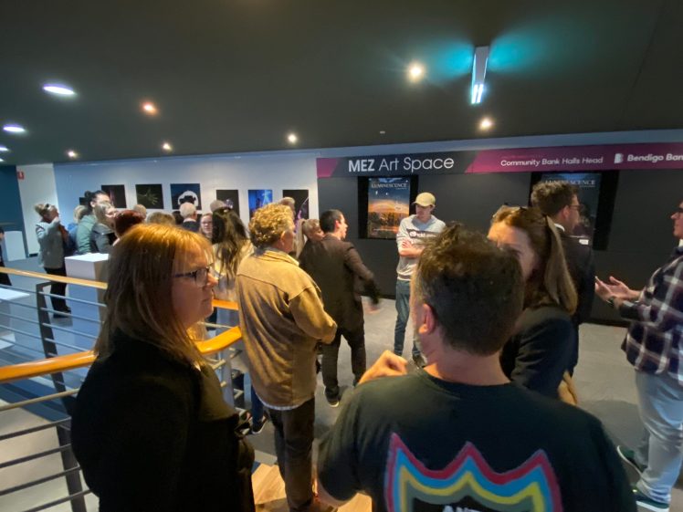 Image of A group of people socialise inside an art gallery called MEZ Art Space. Some view artwork on the walls whilst others converse near a railing in a well-lit, modern interior.