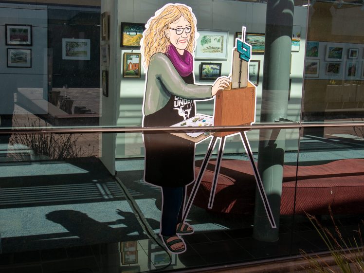 Image of A colourful illustration of a smiling woman painting at an easel is displayed on a glass window, reflecting the outdoor light. Inside, framed artwork hangs on gallery walls.