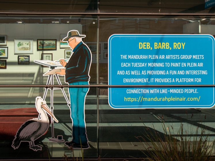Image of A window display shows a painted figure of an artist at an easel with a pelican beside him. A blue sign promotes the Mandurah Plein Air Artists Group and provides website and meeting details. Framed art is visible inside.