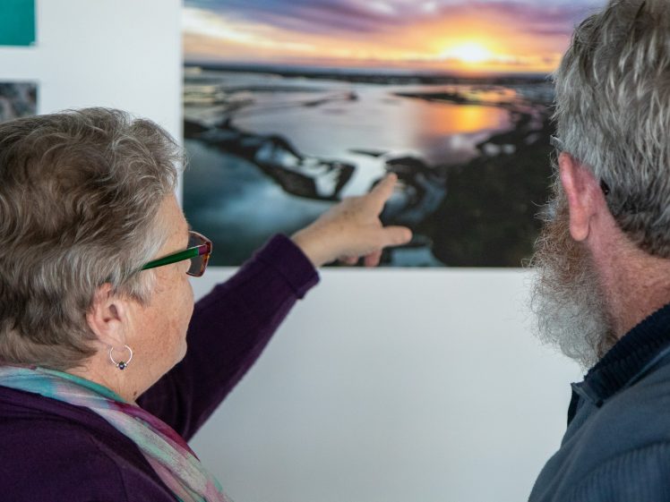 Image of An older woman and man look at a sunset photograph on a wall; the woman is pointing to a feature in the photo. Both are seen from behind, with the man having a grey beard and the woman wearing glasses and earrings.