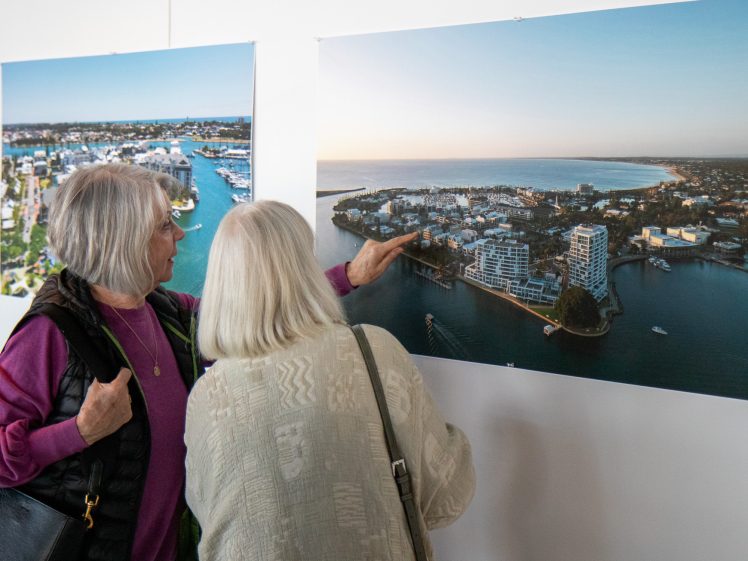 Image of Two women with grey hair look closely at a large aerial photo of a coastal town displayed on a wall. One woman points at a specific spot on the photo as they discuss the image together.