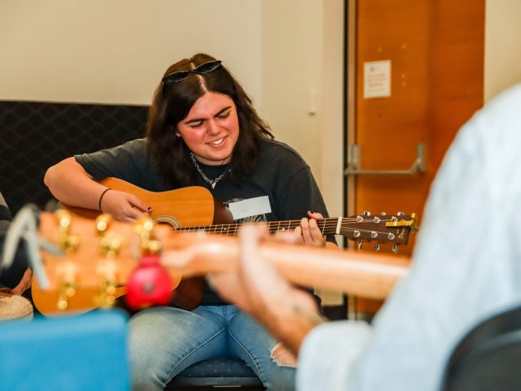 Image of A young woman with dark hair plays an acoustic guitar and smiles with her eyes closed, whilst another person holding a guitar is visible in the foreground. They are indoors, seated in a casual setting.