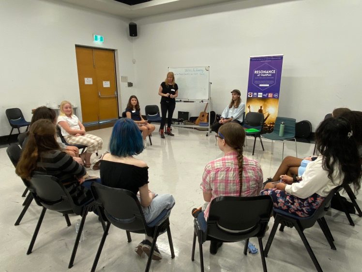 Image of A group of young people sit in a circle in a classroom, listening to a woman standing near a whiteboard and a Resonance Programme banner. A guitar is propped up nearby.
