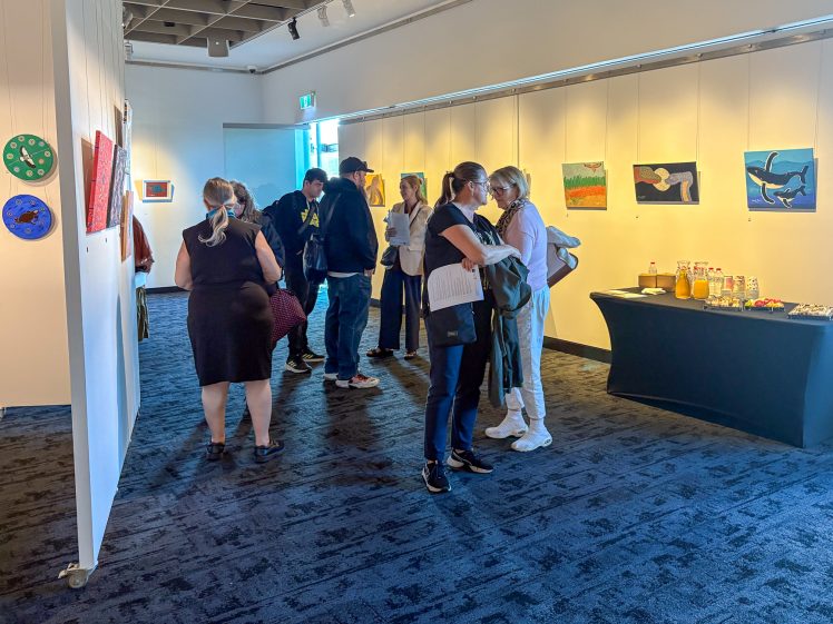Image of People gather in an art gallery with colourful paintings on the walls. Some chat whilst others look at the artwork. A table with drinks and snacks is set up on the right-hand side of the room.