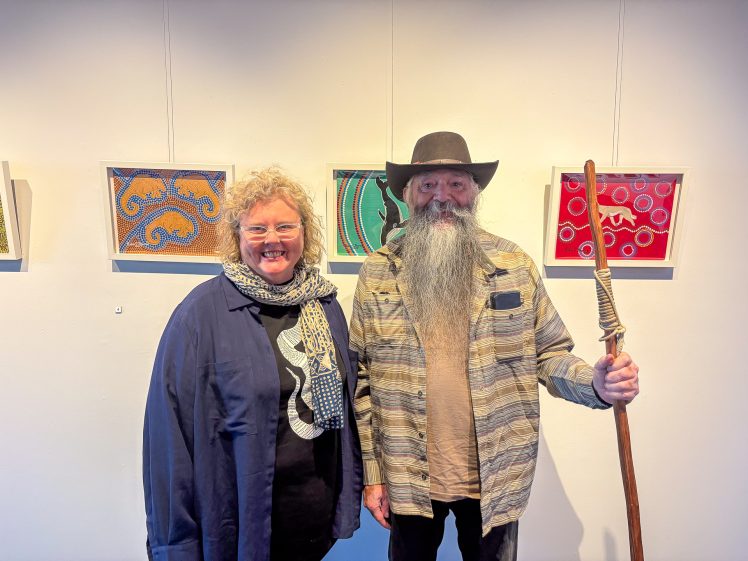 Image of Two people stand indoors in front of colourful abstract artwork. The person on the left has curly blond hair, glasses, a dark jacket, and a patterned scarf. The person on the right has a long grey beard, a black hat, and holds a wooden staff.