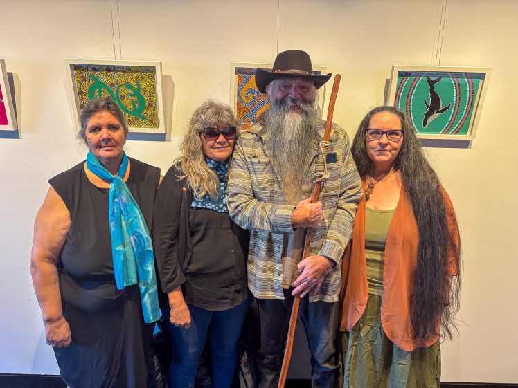 Image of Four adults stand closely together in front of colourful, framed artwork on a white wall. One person holds a wooden staff and wears a wide-brimmed hat. They all look at the camera and appear relaxed and friendly.