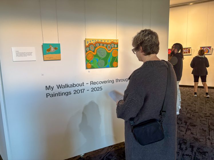 Image of A person with short hair and glasses views colourful paintings on display at an art exhibition titled My Walkabout – Recovering through Paintings 2017–2025. Other visitors look at artwork on adjacent walls.