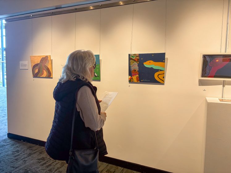 Image of A woman with grey hair stands in an art gallery, holding a piece of paper and viewing colourful paintings featuring abstract and animal designs on a white wall.