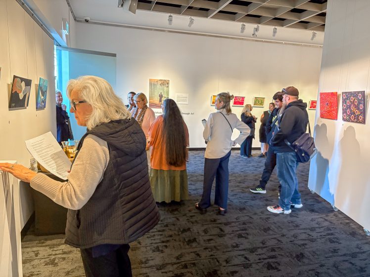 Image of A group of people view artwork displayed on the walls of a brightly lit gallery. Some visitors read information on the walls, whilst others look closely at the paintings. The atmosphere appears calm and focused.