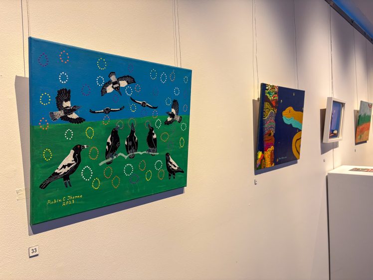 Image of A gallery wall displays colourful paintings. The closest artwork shows black and white birds on green grass and a blue sky, surrounded by dotted circles. Other abstract and landscape paintings are visible further along the wall.