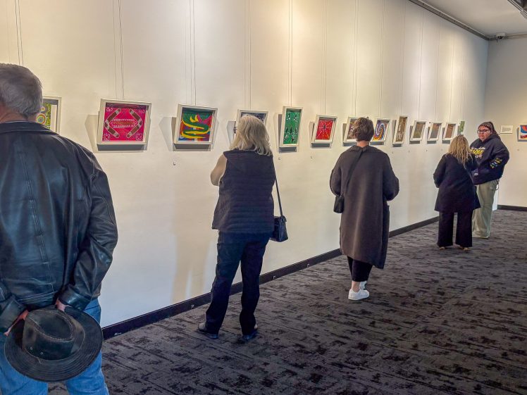 Image of Several people view colourful framed artwork displayed on a white gallery wall. Some stand closely examining the pieces, while others converse in small groups. The room is well-lit with a carpeted floor.