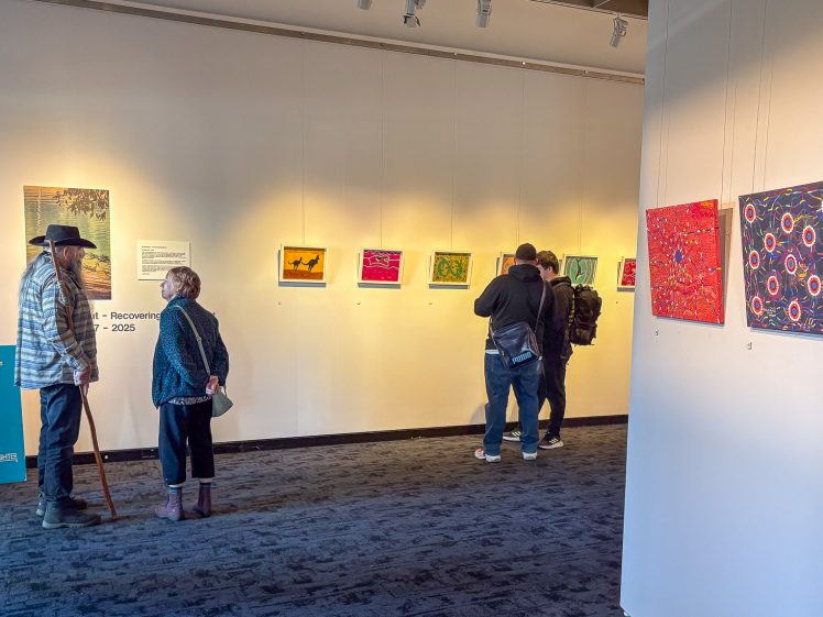 Image of People viewing colourful artworks displayed on white walls in a gallery. Two individuals are conversing on the left, while two others observe paintings on the right. A blue sign offers free LiveLighter recipes. The space is well-lit.