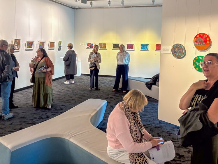Image of A group of people view and discuss artworks displayed on the walls of a modern art gallery; some stand and converse, while others sit on curved white benches. The atmosphere is lively and engaging.
