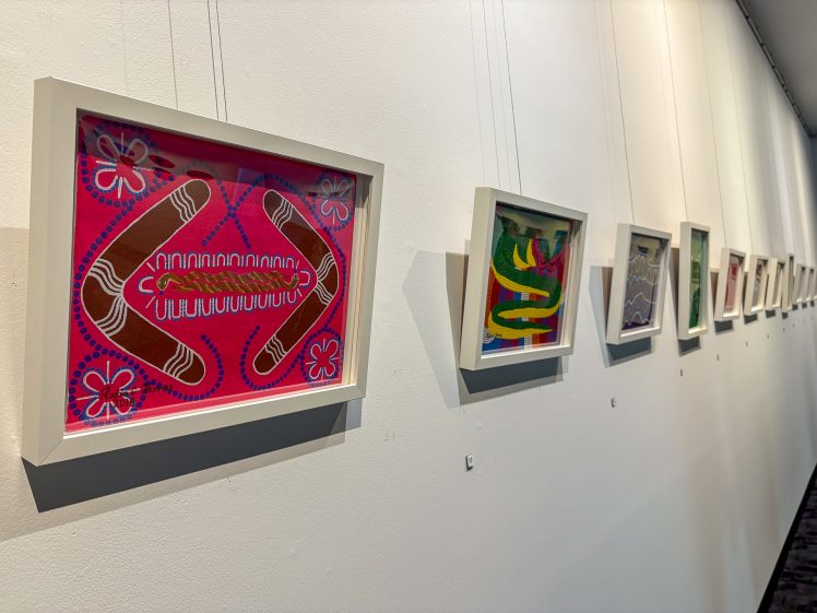 Image of Framed colourful artworks hang in a row on a white wall. The closest piece features boomerang shapes and abstract patterns in red, brown, blue, and pink tones. The other artworks recede into the distance.
