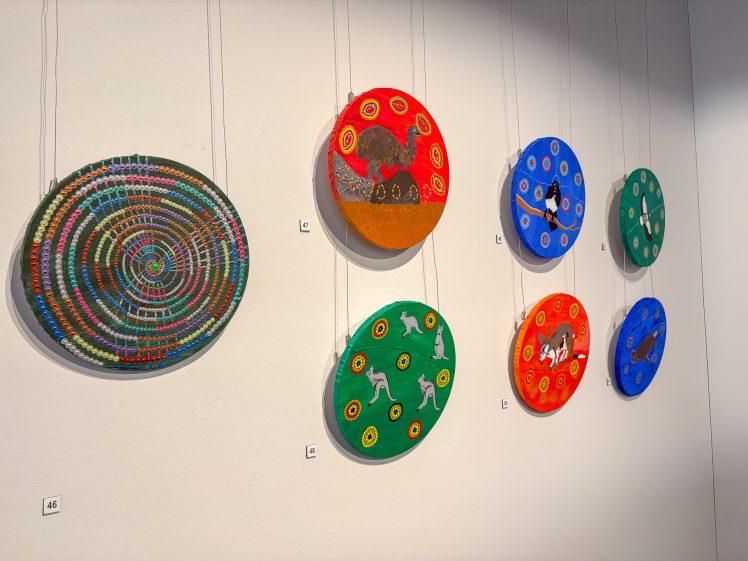 Image of Six colourful round paintings hang on a white wall; five depict animals and patterns, while one features a vibrant spiral dot design. Each artwork is numbered and suspended by visible strings.
