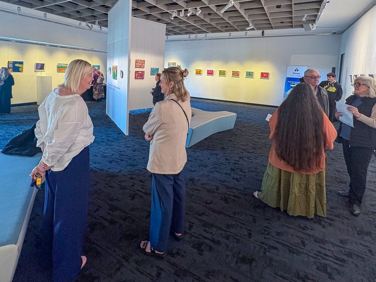 Image of People view and discuss artwork displayed on white walls in a modern, well-lit gallery. Several groups stand and converse whilst others observe the art. The space has dark flooring and white benches.