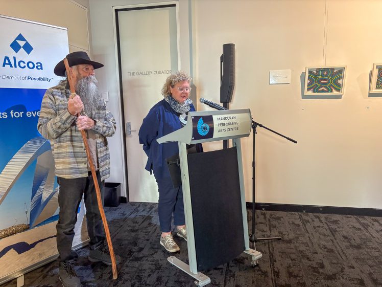 Image of A woman speaks at a lectern labelled Mandurah Performing Arts Centre whilst a man in a hat with a long beard and walking stick stands beside her. Artworks and an Alcoa sponsorship banner are visible in the background.