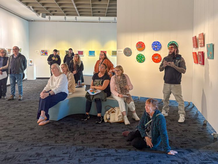 Image of A group of people sit and stand in a modern art gallery, observing colourful artwork displayed on white walls. Some viewers sit on benches, while others stand or sit on the floor. The atmosphere appears calm and attentive.