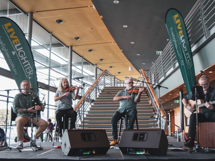 Image of Four musicians perform on a stage indoors, with two playing violins, one playing a flute, and one on guitar. They sit in front of stairs and are flanked by two green LiveLighter banners.