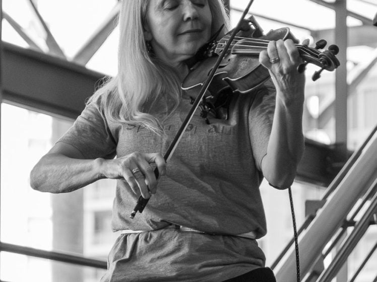 Image of A woman with long hair plays the violin while seated, eyes closed in concentration, next to a microphone in a modern indoor space with large windows and metal beams. The image is in black and white.
