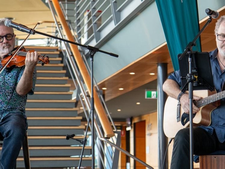 Image of Two men perform indoors beside a staircase; one plays a violin while seated and the other plays an acoustic guitar and sings into a microphone. Both wear glasses and casual clothing.