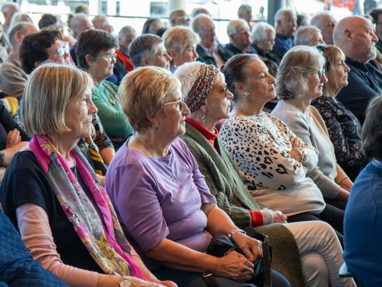 Image of A large group of older adults sits closely together in rows, attentively listening to something out of view. Many appear engaged and focused, with neutral or thoughtful expressions.