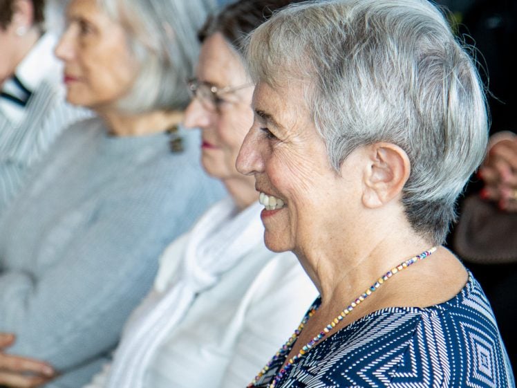Image of A group of older women sit in rows, attentively listening and smiling during an indoor event. The focus is on a woman in the foreground wearing a blue patterned top and a beaded necklace.