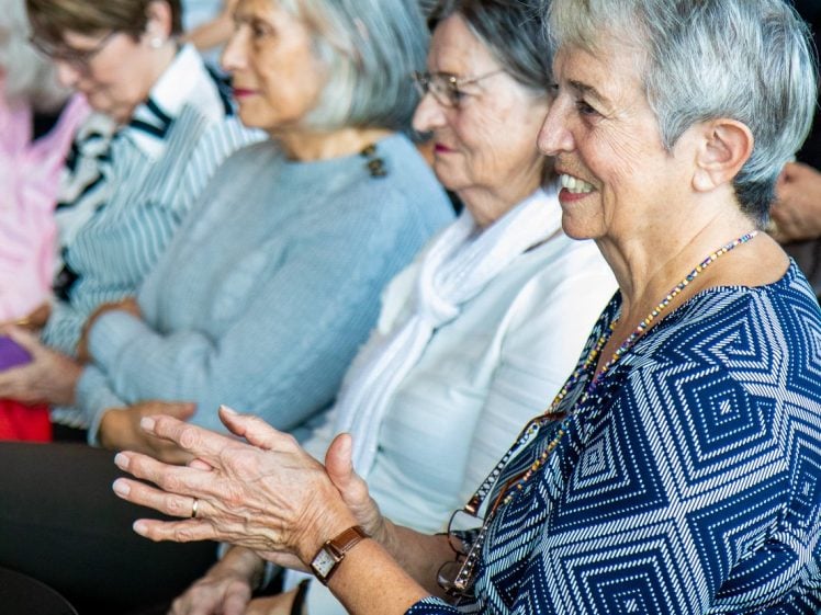 Image of A group of older women sit closely together in an audience, smiling and applauding while attentively watching an event or presentation.