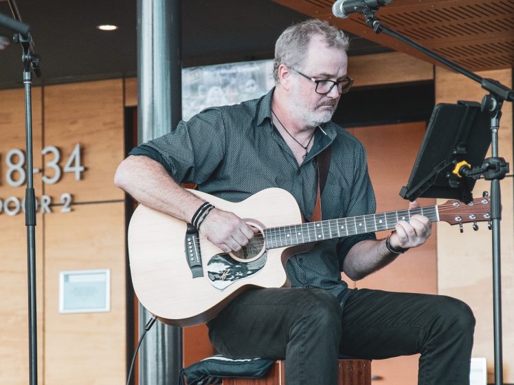 Image of A man with glasses plays an acoustic guitar on stage. He is seated, wearing a dark shirt and jeans, and focussing on his performance. A microphone and tablet are positioned in front of him.
