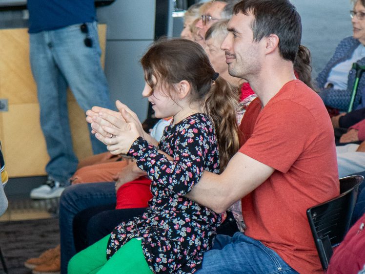 Image of A man in a red shirt sits with a young girl in a floral dress on his lap. They are in an audience, and the girl is clapping whilst others watch attentively.