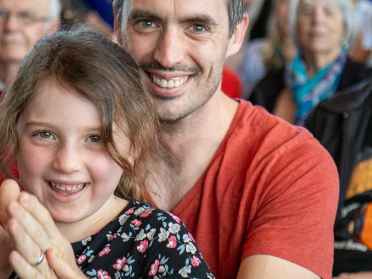 Image of A smiling man in a red shirt sits closely behind a young girl in a floral dress, holding her hands. Both are looking at the camera, with a blurred audience seated in the background.