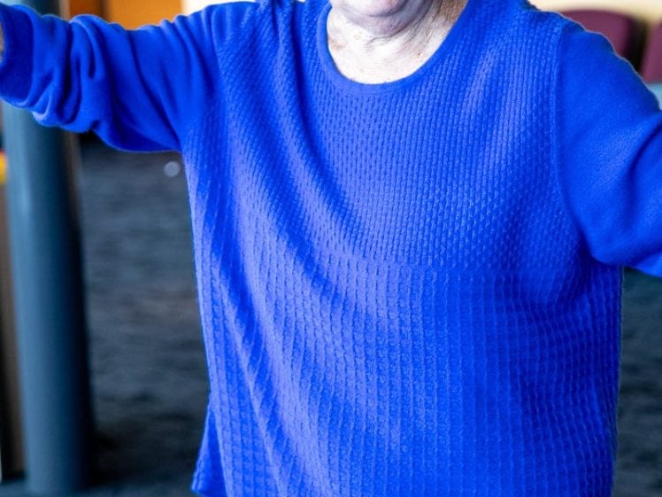 Image of An older woman with short white hair, wearing a bright blue textured jumper and earrings, stands indoors with her arms outstretched, appearing to speak or gesture expressively.