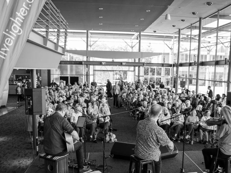 Image of A black and white photo of musicians performing on a stage in front of a large seated audience in a modern glass-walled venue. The view is from behind the band, looking towards the crowd.