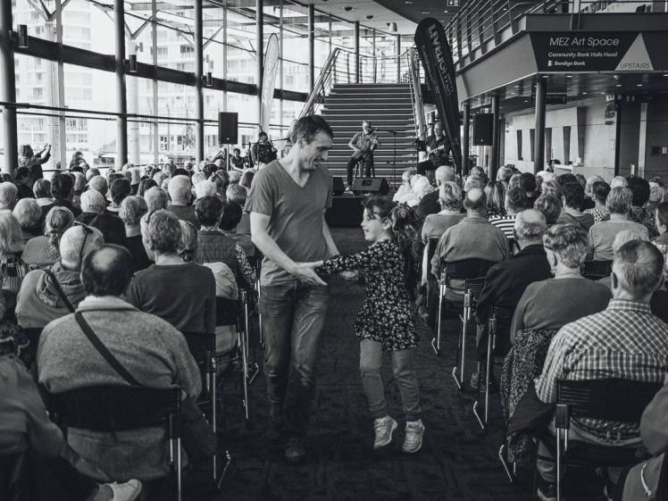 Image of A man and a young girl dance in the aisle among a seated audience watching a live performance on stage inside a modern, glass-walled building. The scene is lively and filled with natural light.