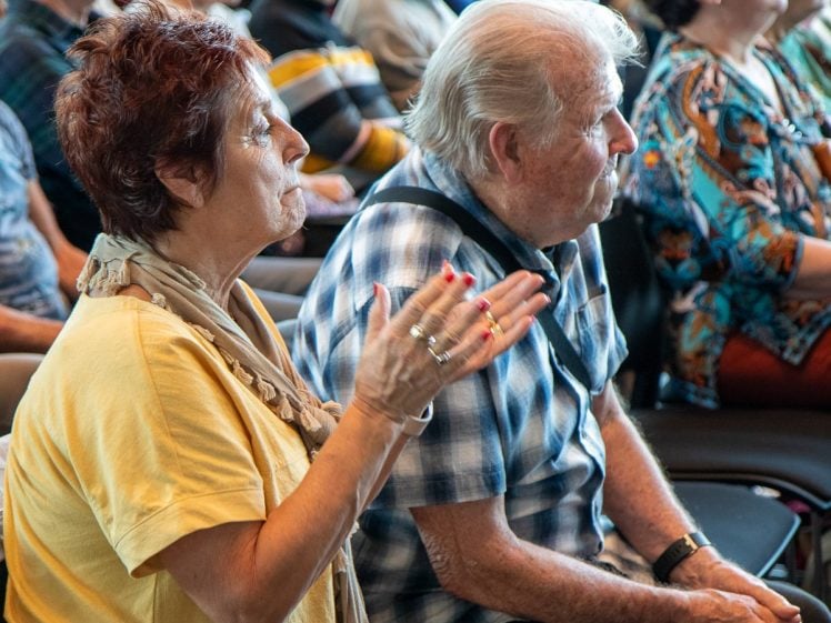 Image of An older woman wearing a yellow top gestures with her hands while seated next to an older man in a checked shirt at an indoor gathering with other people in the background.