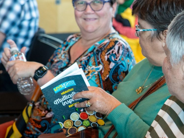 Image of Three people sit together. One person holds a booklet titled Healthy Multicultural Inspired Recipes whilst another smiles at the camera. The group appears to be at an indoor event.